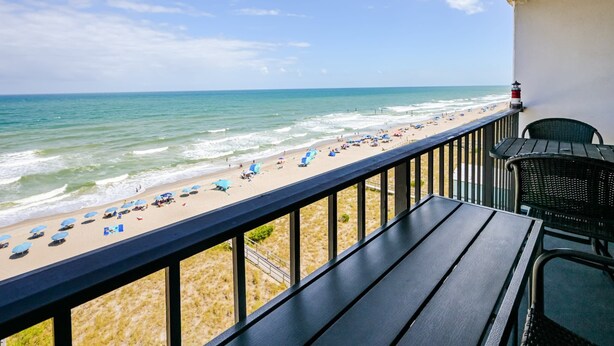 A balcony with a view of a beach, a lighthouse, and a chair. - Octopus Garden: 1BR, King Bed, Indoor/Outdoor Pool (Carolina Beach)