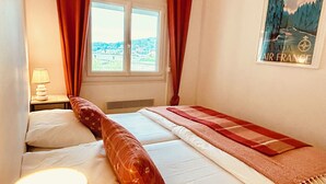 Hotellihuone, jossa on sänky, ikkuna, jossa on verhot, lamppu ja taulu seinällä.