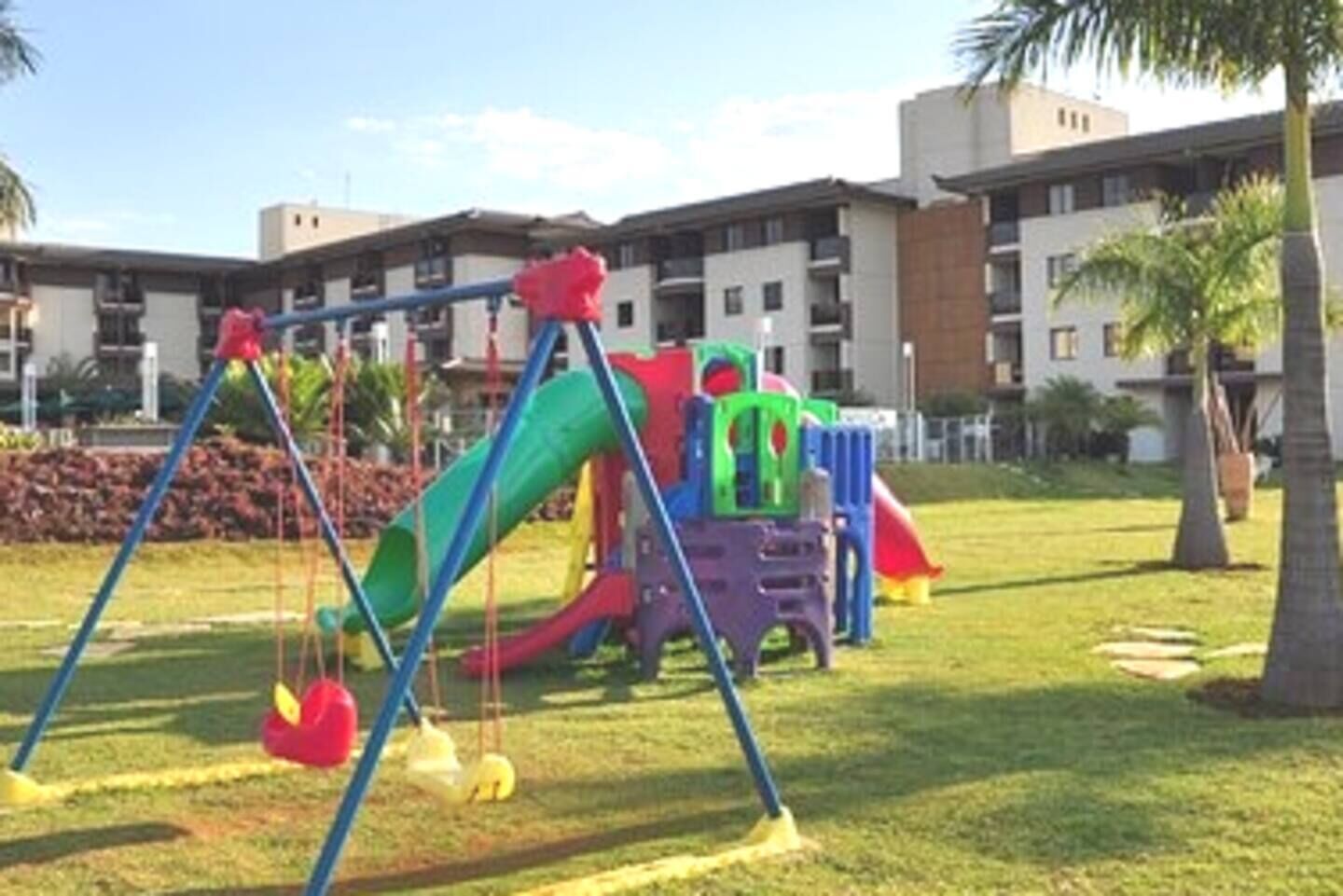 A playground with swings and slides, surrounded by a grassy area and palm trees, with apartment buildings in the background.