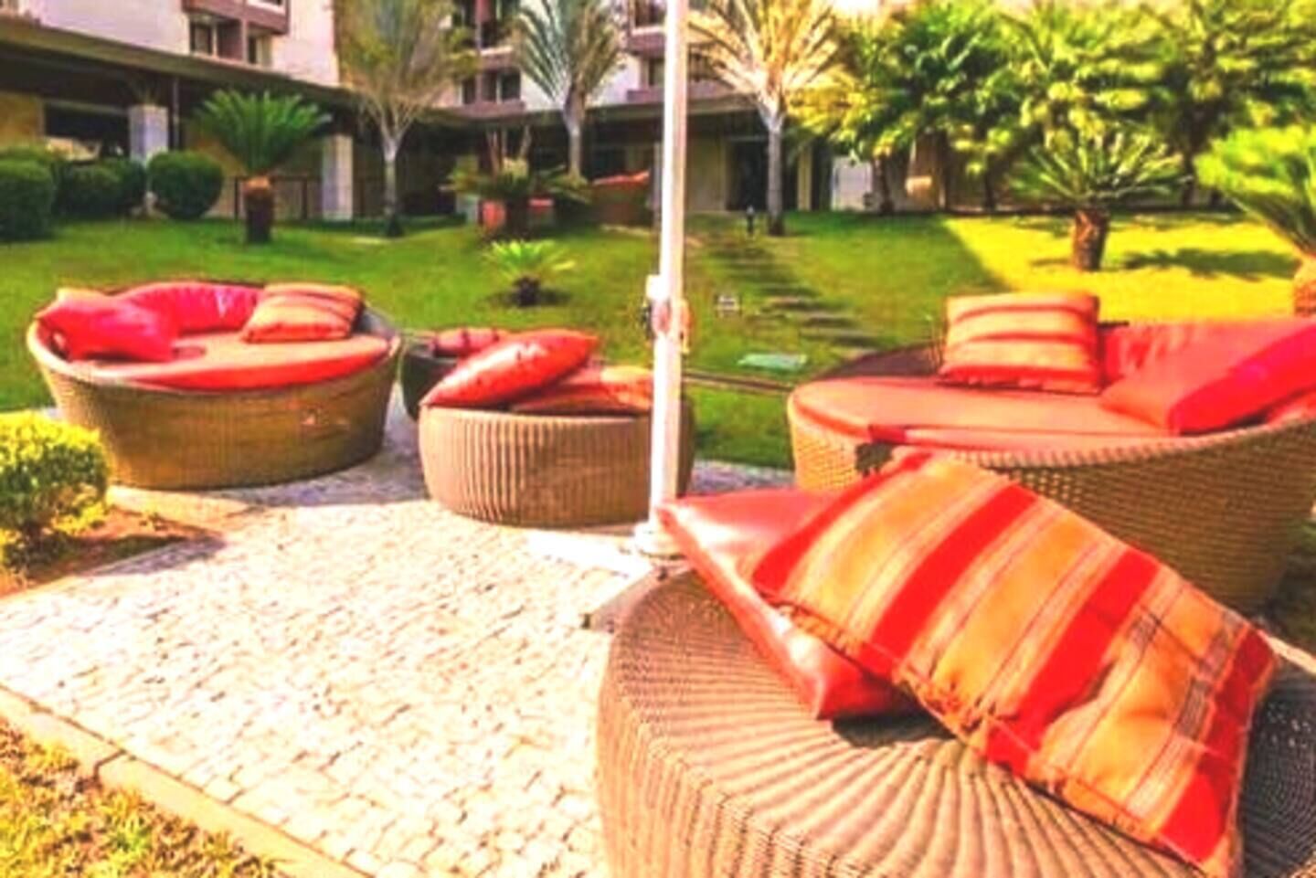 A courtyard with wicker furniture and red cushions, surrounded by greenery and a building in the background.