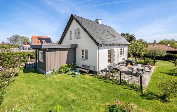 Una casa de dos pisos con techo inclinado, jardín cercado y patio con mobiliario exterior.