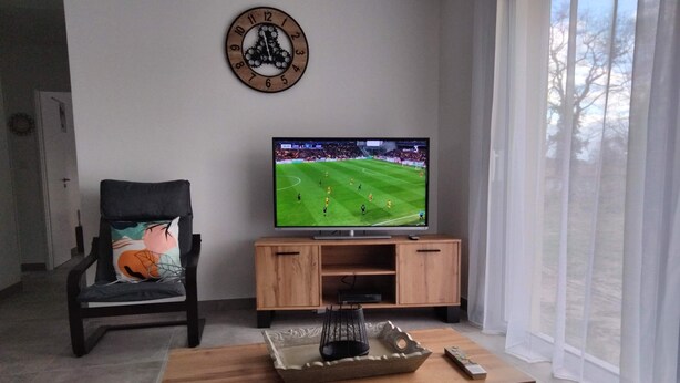 A living room with a television on a wooden stand, a chair with a cushion, and a clock on the wall. - Gite DU Vieux Chene (Reignac)