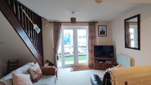 A living room with a staircase, a television, and a view of boats through the glass doors. - Cosy and comfortable Riverside Cottage in Wroxham, the Gateway to the Broads (Wroxham)
