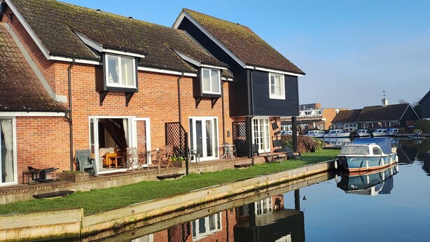 A canal-side property with a brick house, a boat docked on the water, and a small boat in the foreground. - Cosy and comfortable Riverside Cottage in Wroxham, the Gateway to the Broads (Wroxham)