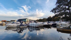 A marina with several boats docked, a large building with a blue roof, and a calm water surface reflecting the sky. - Cosy and comfortable Riverside Cottage in Wroxham, the Gateway to the Broads (Wroxham)
