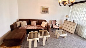 Un salon avec un canapé d’angle marron, deux petites tables d’appoint blanches, un fauteuil à motifs, une commode blanche et un lustre.