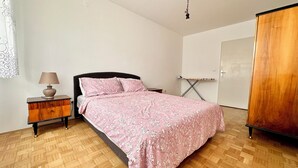 Une chambre avec un plancher en bois, un lit recouvert d’une couvre-lit à motifs floraux, une table de chevet avec une lampe et une armoire.