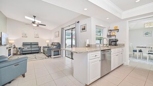 A modern kitchen with a central island, a dining area with a table and chairs, and a living room with sofas and a TV. - Custom Pool! Tiki Bar | Private Yard ~ Enjoy SWFL (Fort Myers)