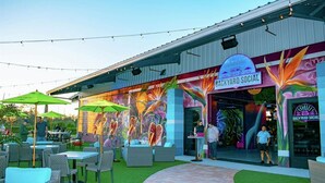 An outdoor seating area with wicker furniture and umbrellas, a colorful mural on the building, and a sign reading "BACKYARD SOCIAL". - Custom Pool! Tiki Bar | Private Yard ~ Enjoy SWFL (Fort Myers)