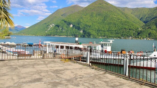 A ferry dock with a boat, a concrete platform, and a mountain in the background. - The Painter's Nest + Lake Garden (Cima)