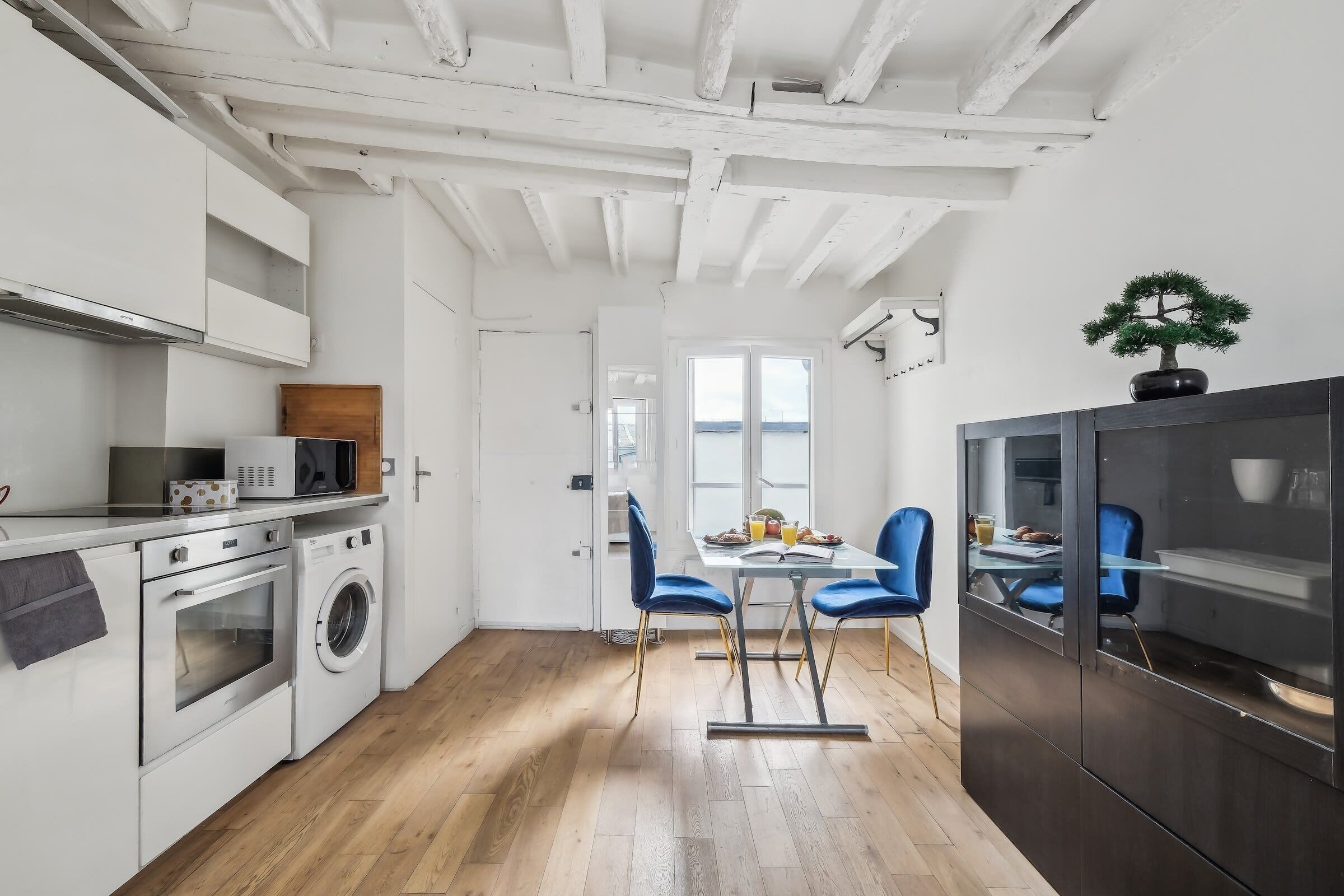 A modern kitchen with white cabinetry, a built-in oven, and a washing machine. There is a dining area with a table and chairs, and a view of the outside through a window.