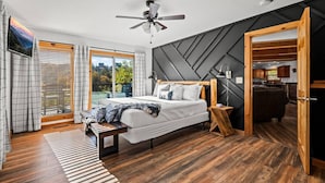 Un dormitorio moderno con una cama grande, suelo de madera, un ventilador de techo y vistas al exterior a través de un ventanal corredero.