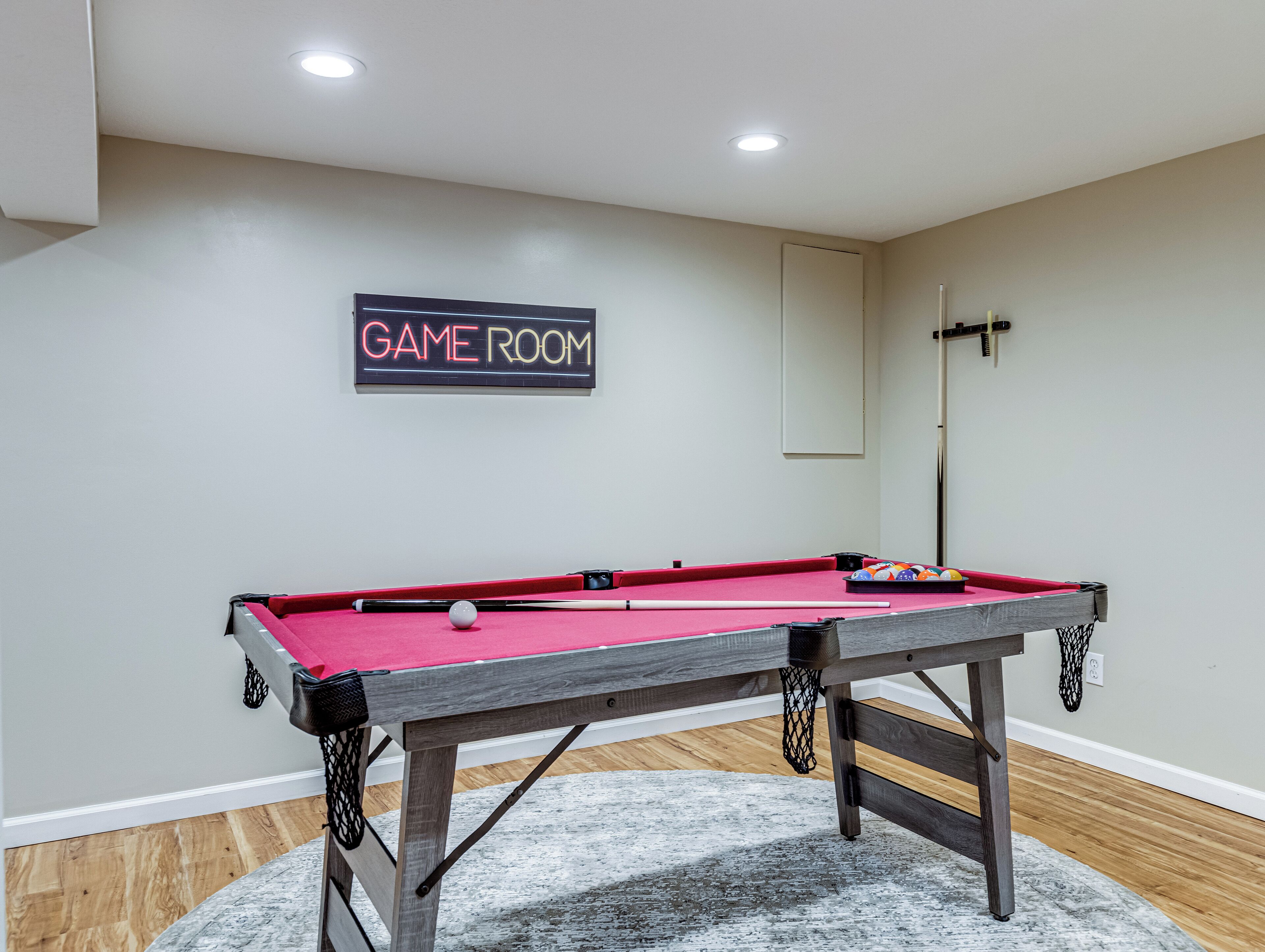 A pool table with a 'GAME ROOM' sign, a mirror, and a basketball hoop in a room with wooden flooring and a gray carpet.