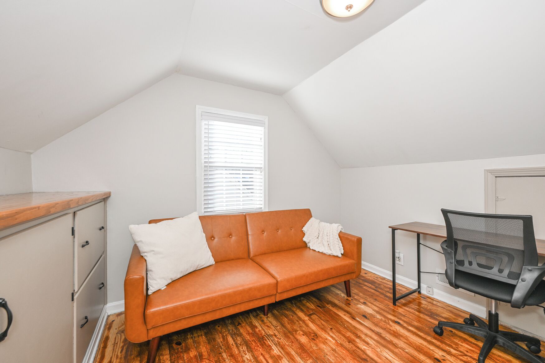A small, well-lit room with a wooden desk, a leather sofa, and a black office chair.