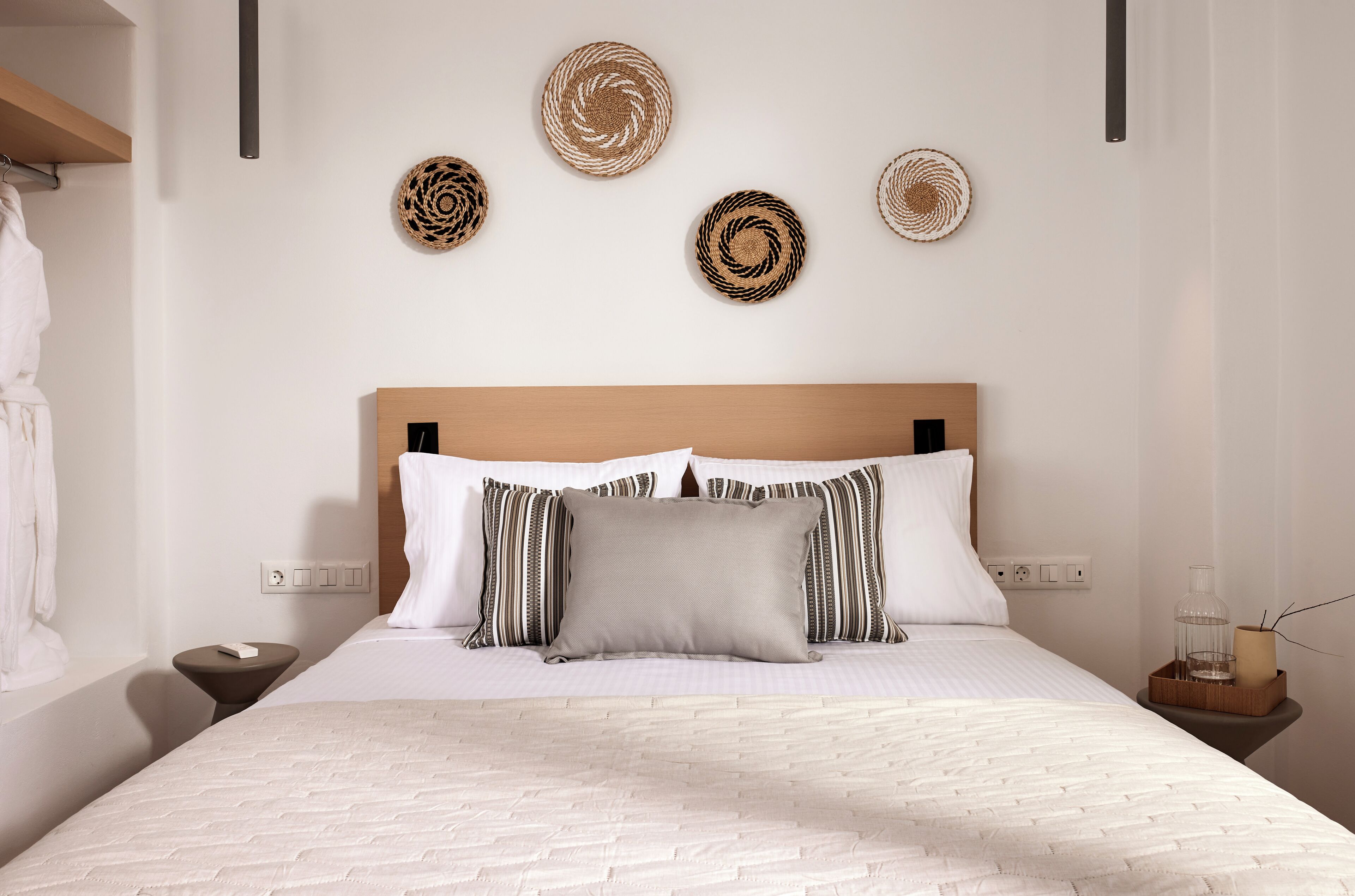 Ein ordentlich bezogenes Bett mit Holz Kopfteil, ein Nachttisch mit Lampe und an der Wand befestigte gewebte Körbe.