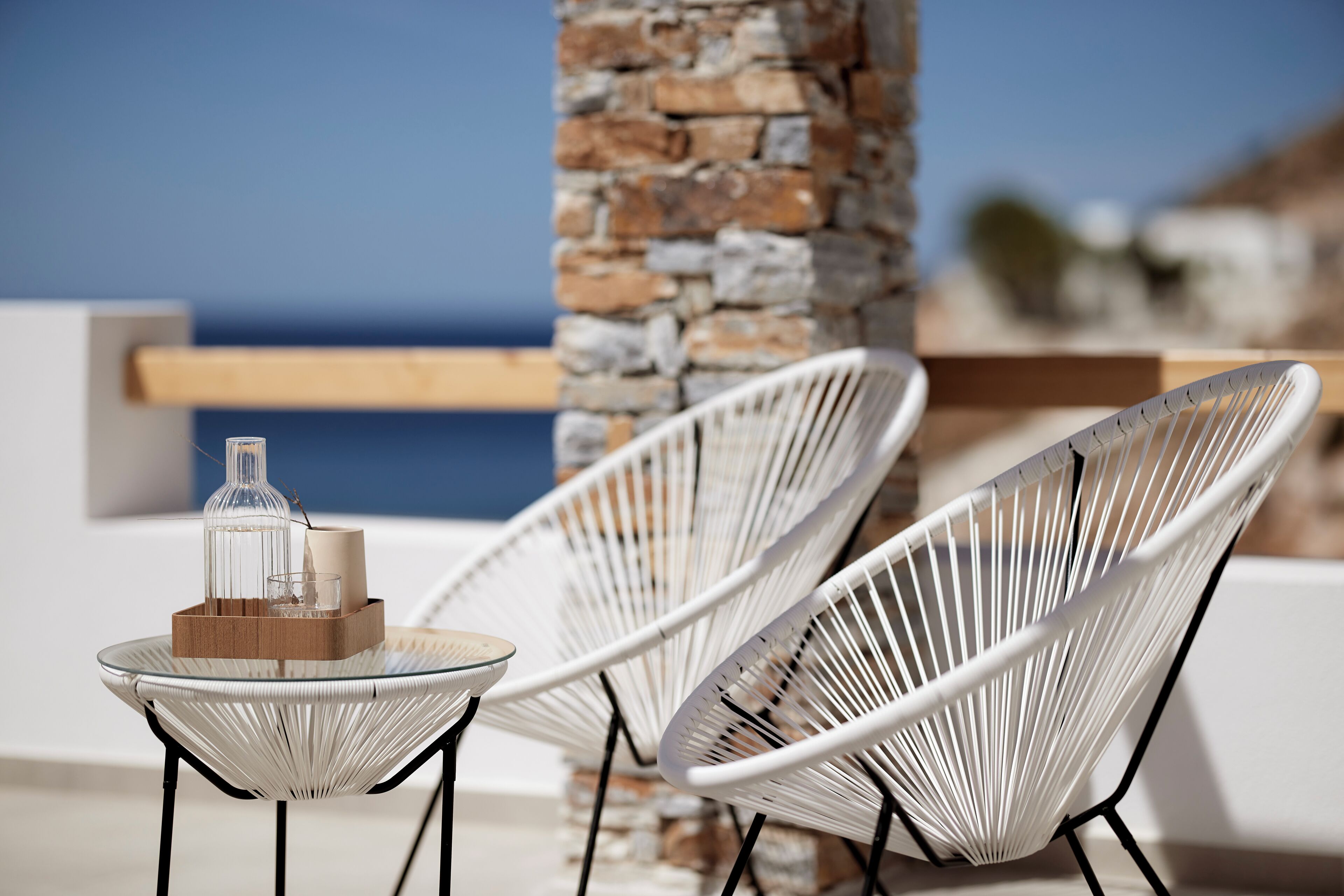A glass table with a bottle and a glass, two white wire chairs, and a stone pillar.