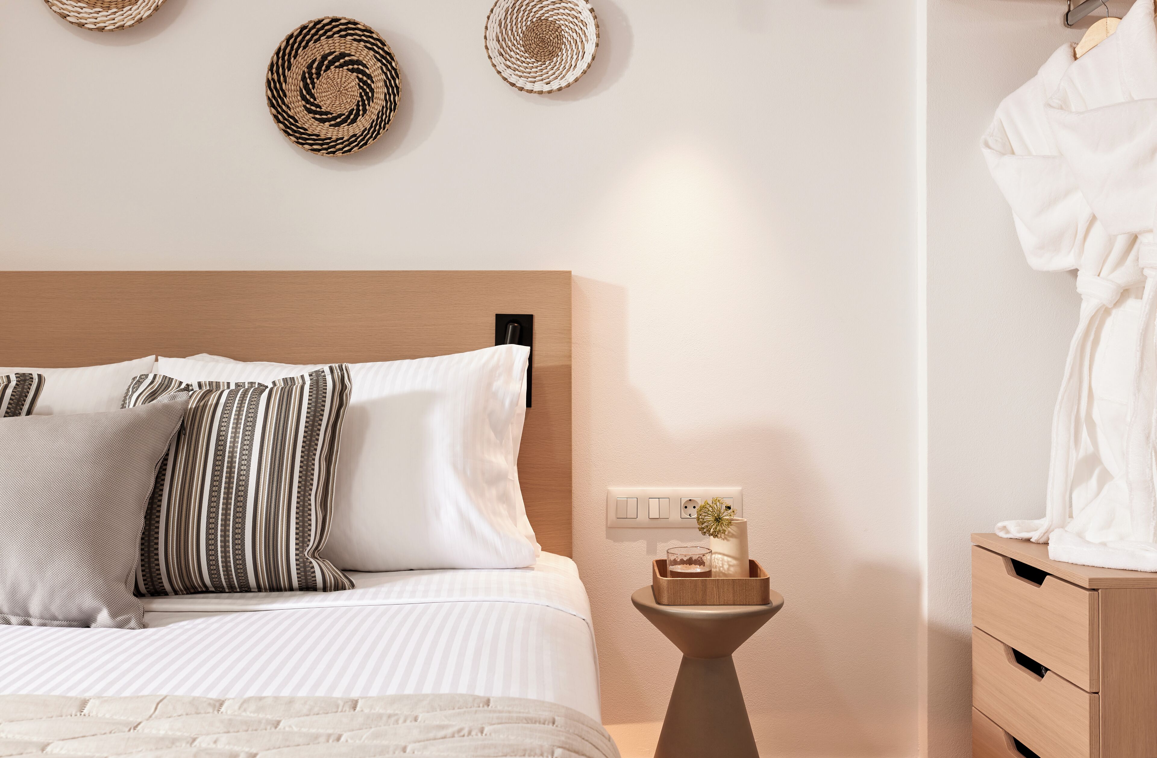 Un dormitorio ordenado con cama, almohadas, una mesita de noche con lámpara y un tocador de madera.