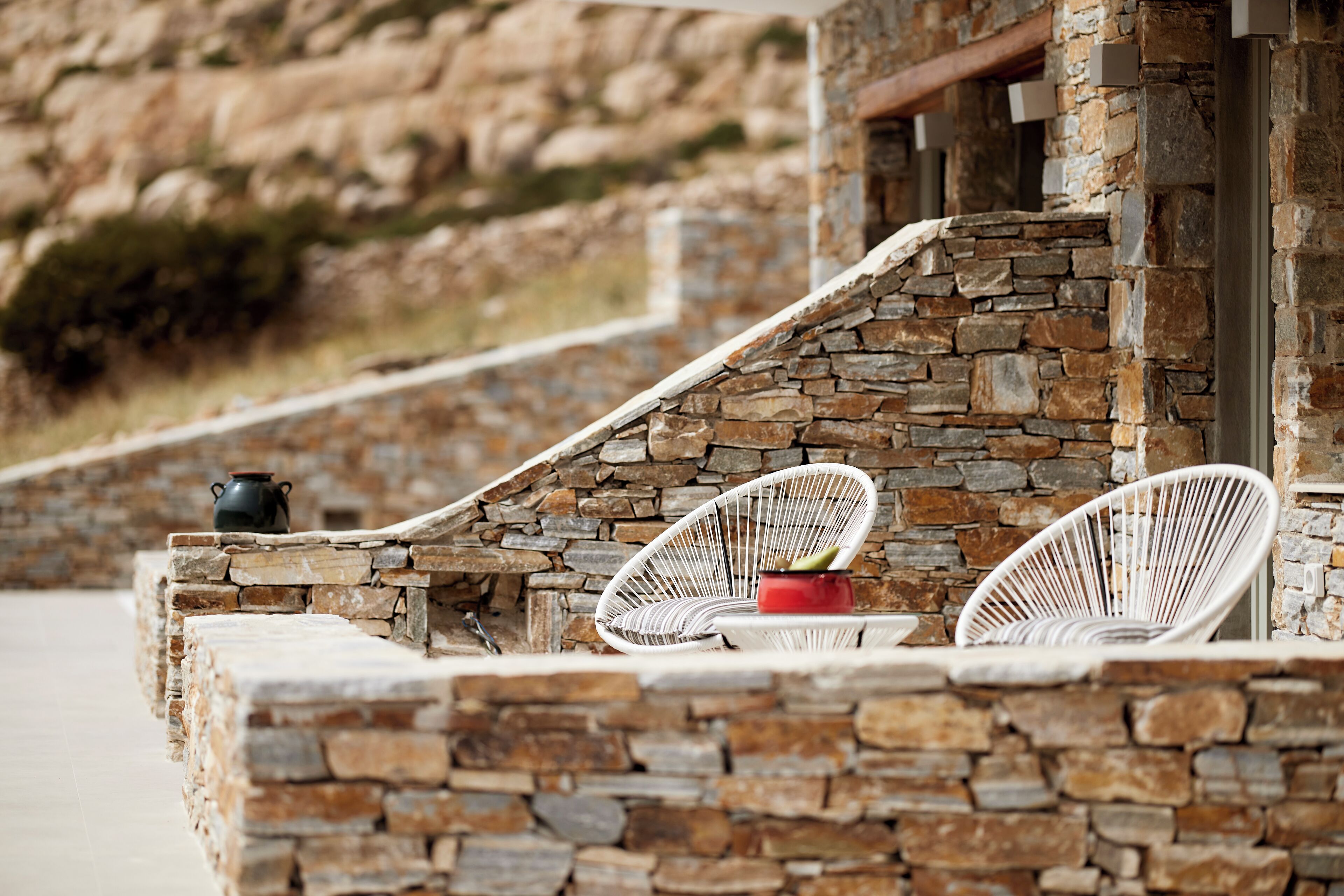 A stone-built outdoor seating area with wicker chairs and a small table, overlooking a rocky landscape.
