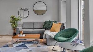 Ein modernes Wohnzimmer mit grauem Sofa, einem grünen Sessel, einem Holztisch und einem gemusterten Teppich. Es befinden sich zwei Wandspiegel und ein Blumentopf dort.