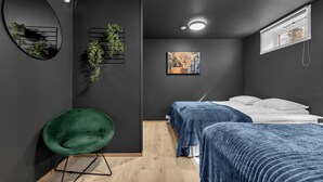 Ein modernes Schlafzimmer mit dunkler Wand, einem grünen Sessel, einem Bett mit blauer Bettdecke, einem Gemälde und einem Fenster mit Blick.