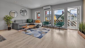 Ein modernes Wohnzimmer mit einem grauen Sofa, zwei grünen Sesseln, einem Couchtisch und einem großen Fenster mit Blick auf farbenfrohe Gebäude.