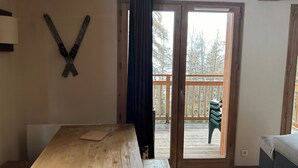 Una mesa de madera con un tapete de corcho, una puerta de madera con una cortina y vistas a un balcón con una silla.