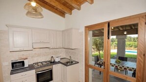 A kitchen with white cabinets, a microwave, stove, and sink. There's a sliding glass door leading to an outdoor patio with a table and chairs. - Villetta Saeprus (Muravera)