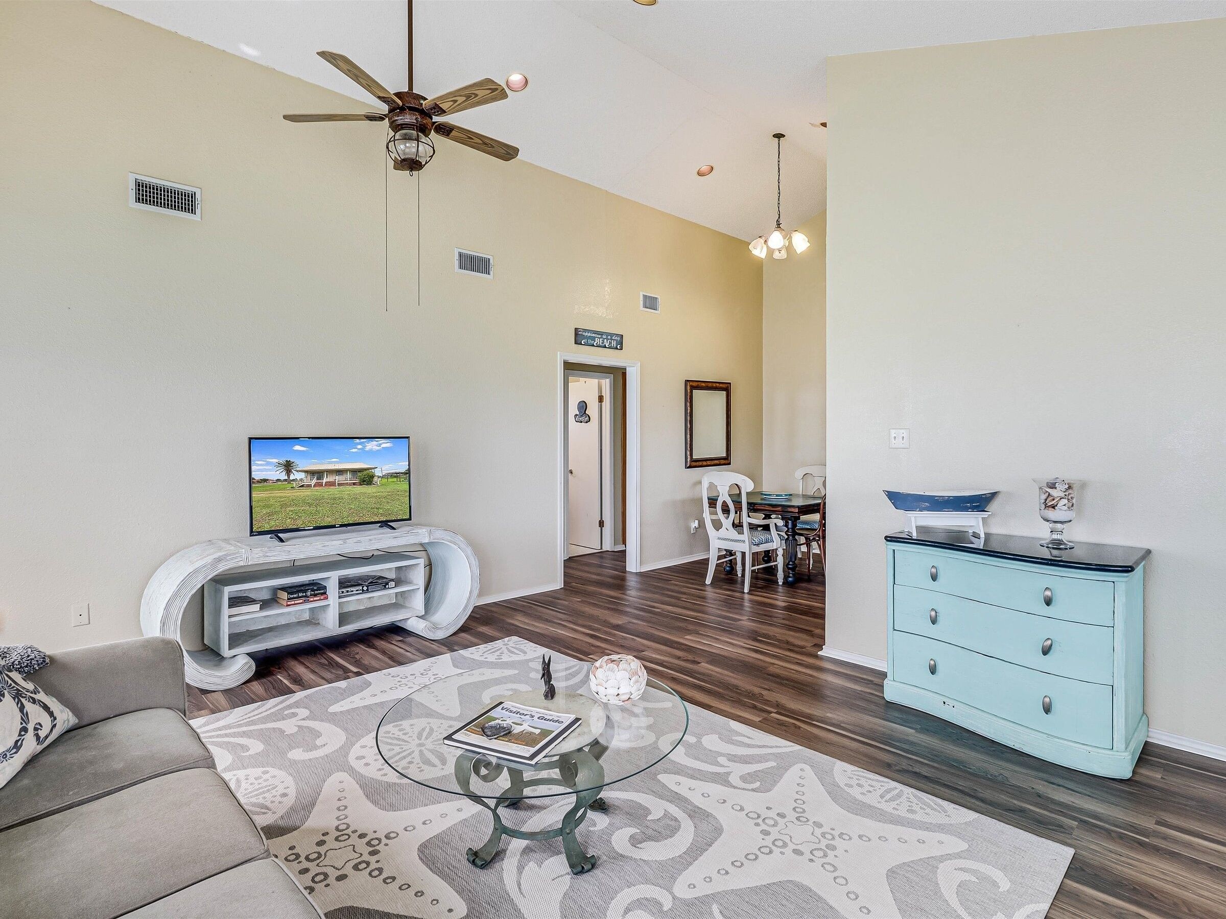 A living room with a TV, a ceiling fan, a dining area with a table and chairs, and a light blue dresser.