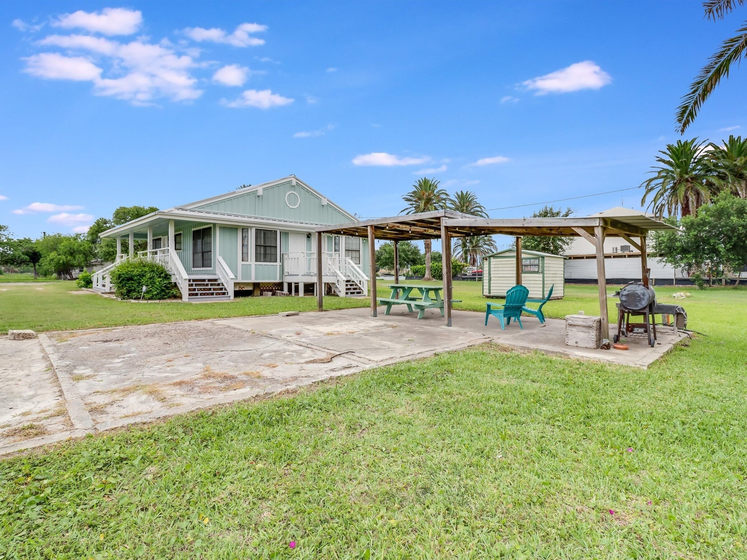 A house with a porch, a covered patio area with a picnic table and chairs, and a small shed in the background.
