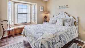 A bedroom with a large bed, a window with blinds, a bedside table with a lamp, and a wall-mounted sign that says "RELAX".