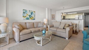 Un salón moderno con dos sofás beige, una mesa de centro de vidrio y una cocina al fondo.