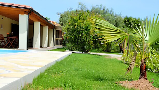 A poolside area with a clear view of the pool, a covered patio with chairs, and a lush green lawn. - An oasis of tranquility near the beautiful beaches of Sarrabus. (Muravera)