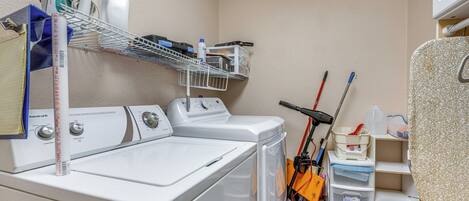 A laundry area with a washing machine, dryer, ironing board, and shelves with various items.
