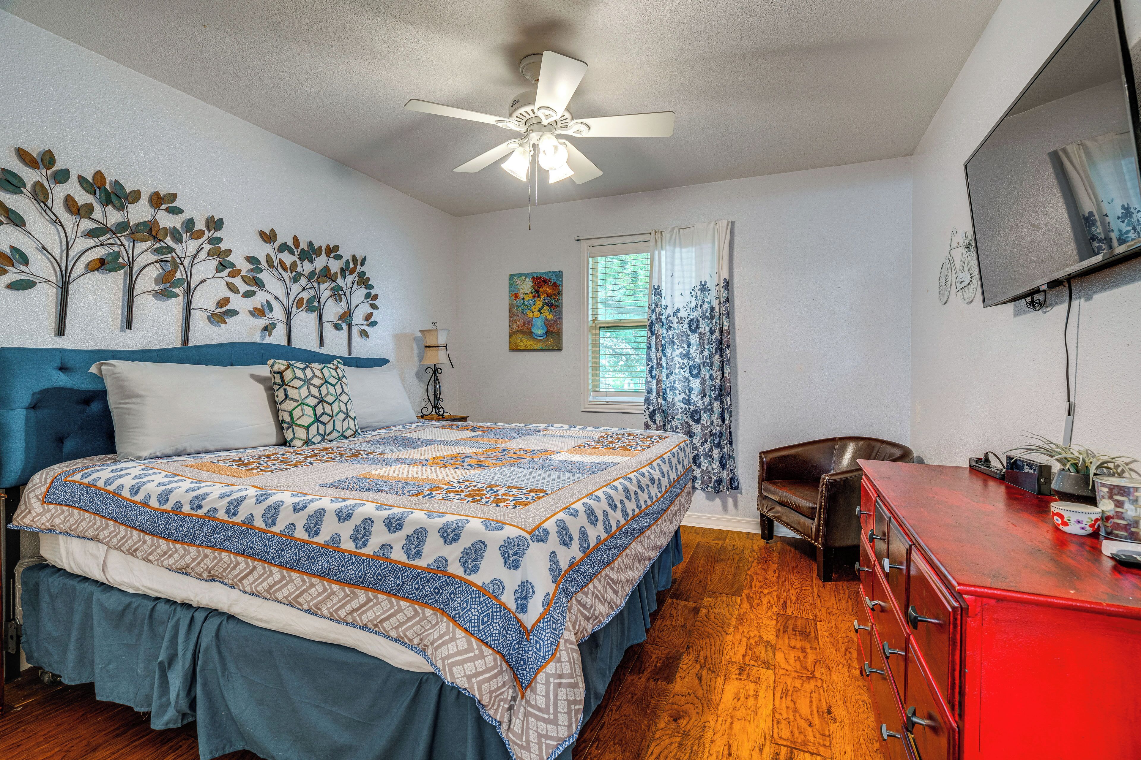 A bedroom with a bed, a dresser, a chair, a ceiling fan, a window with curtains, and wall art.