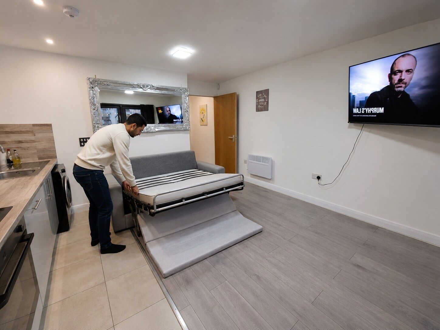 A modern room with a bed, a television, a mirror, and a kitchen area.