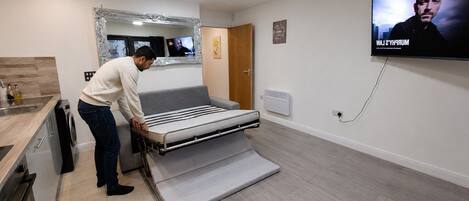 A modern room with a bed, a television, a mirror, and a kitchen area.