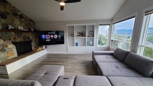 A modern living room with a stone fireplace, a flat-screen TV, grey sofas, and a large window offering a view of the landscape.