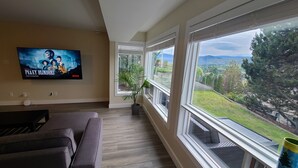 A living room with a large TV displaying 'Peaky Blinders', a potted plant, and a view of a green landscape through large windows.