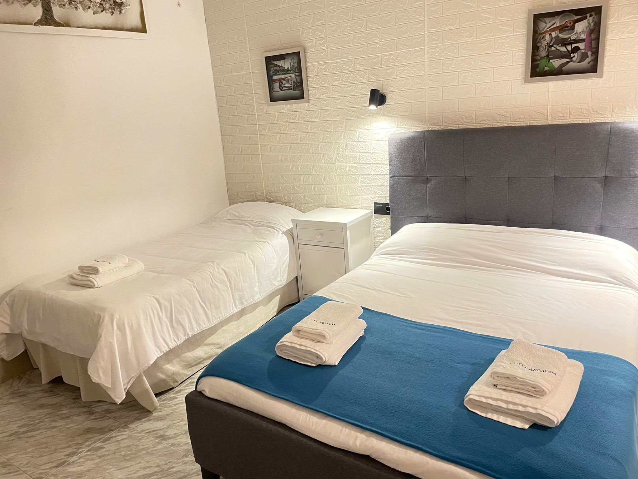 Habitación de hotel con dos camas individuales, una cama grande, una mesita de noche y una lámpara fijada a la pared.