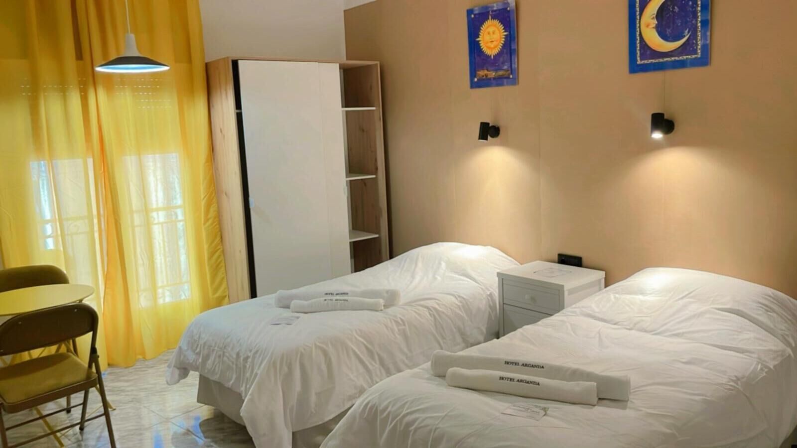 A hotel room with two beds, a desk, and a chair. There are two wall-mounted nightstands and a wardrobe.