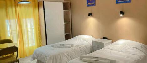 A hotel room with two beds, a desk, and a chair. There are two wall-mounted nightstands and a wardrobe.