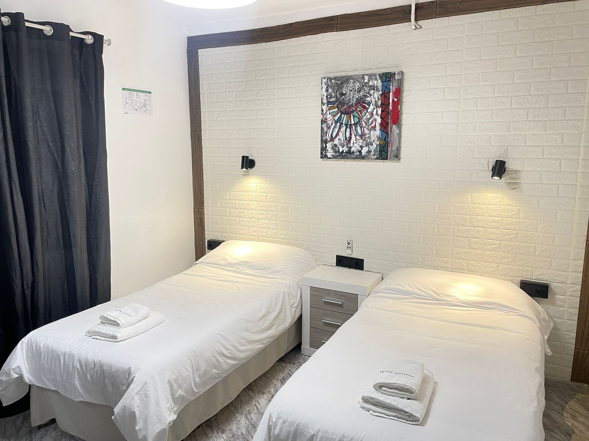 A room with two single beds, a small bedside table, a wall-mounted artwork, and a curtain.
