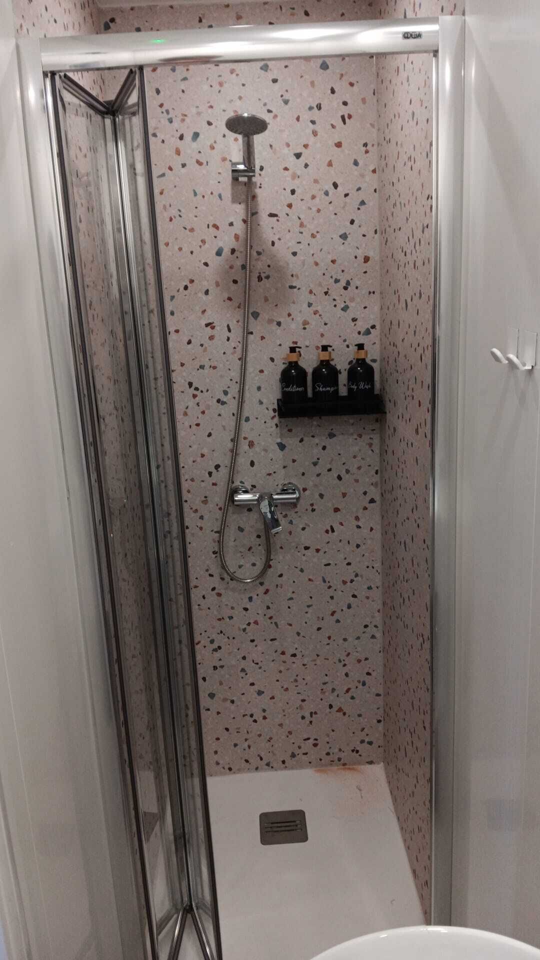 A shower with a glass door, a handheld showerhead, and a shelf with toiletries.