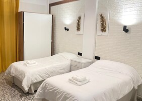 Habitación de hotel con dos camas individuales, un armario, una mesita de noche pequeña y una lámpara fijada a la pared.