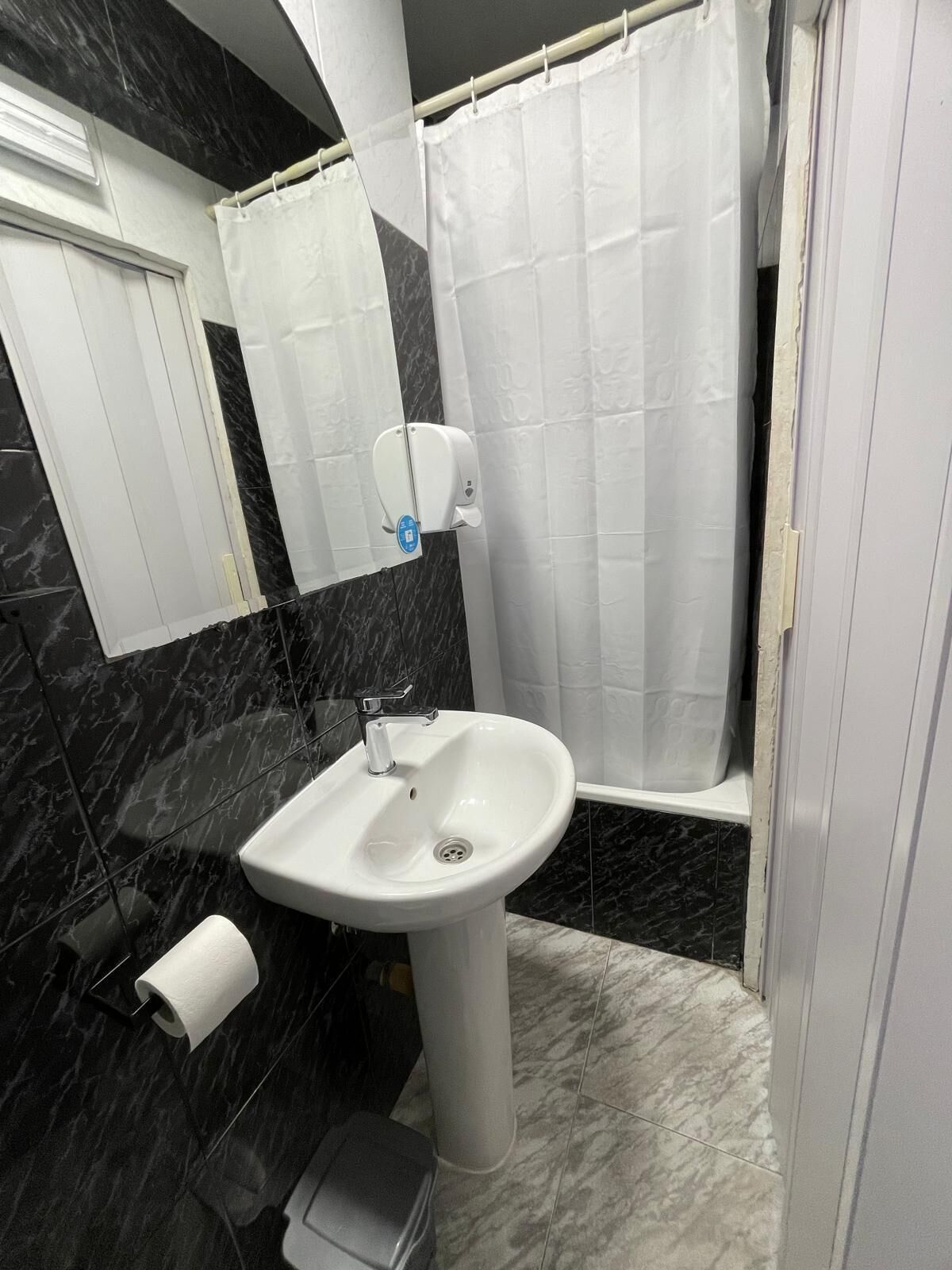 Un bagno con un lavandino bianco su piedistallo, uno specchio, un dispenser per asciugamani, un porta carta igienica e una tenda da doccia.