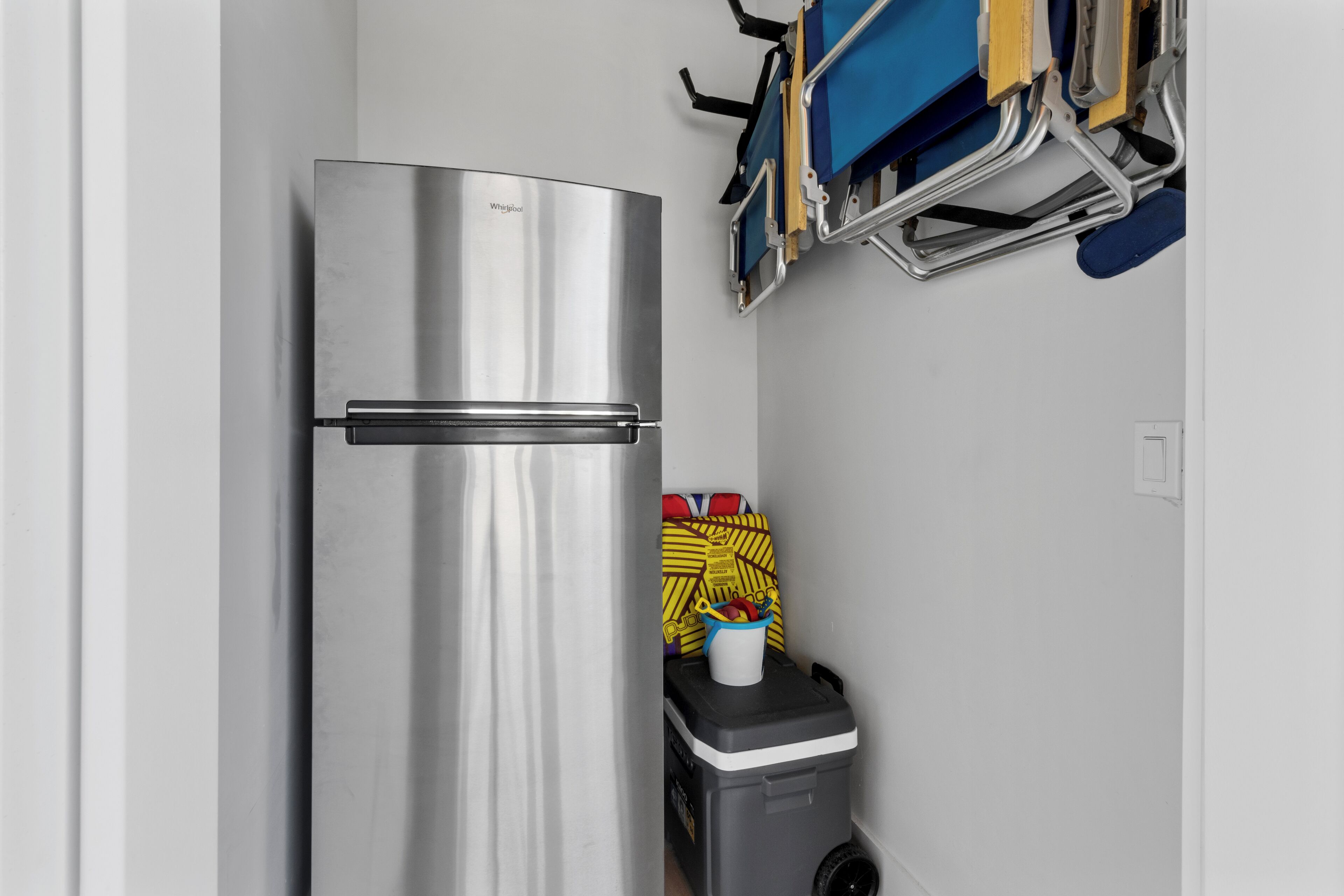 A compact kitchen with a stainless steel refrigerator, a small trash bin, and a blue chair with a yellow cushion.