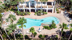 Eine Vogelperspektive zeigt einen luxuriösen Urlaubsort mit einem zentralen Pool, zahlreichen Balkonen und Palmen.