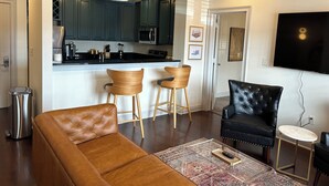 Ein modernes Wohnzimmer mit einem braunen Ledersofa, zwei Holzbarhocker, einem schwarzen Ledersessel, einem Glastisch und einem flachbildfernseher an der Wand.