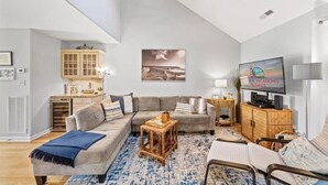 Un salon spacieux avec un canapé gris, une table basse et un téléviseur. On y trouve également un meuble en bois et une lampe sur le côté.