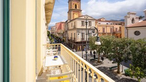 Un balcón con mesa y sillas, con vista a una calle con edificios y una torre del reloj.